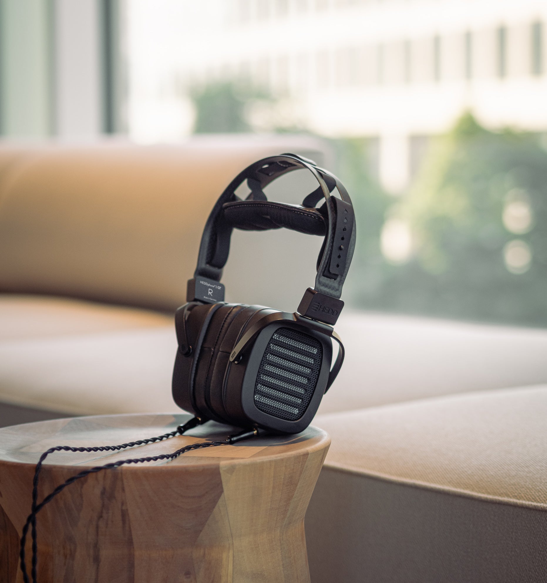 A pair of over-ear, open-back HEDDphone TWO GT headphones resting on a wooden surface with a blurred background featuring a cushioned chair and large windows.