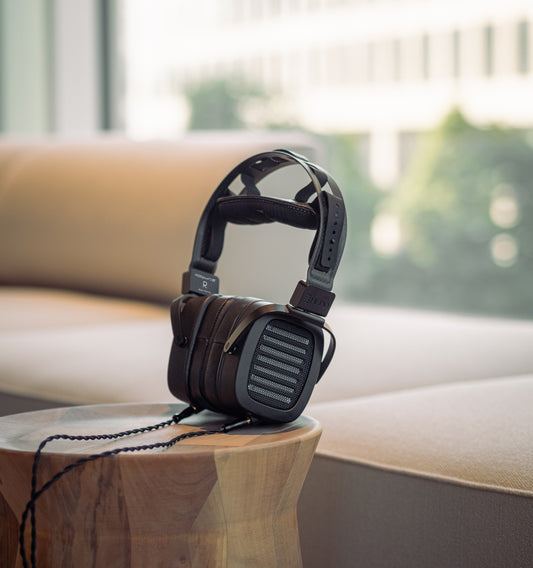 A pair of over-ear, open-back HEDDphone TWO GT headphones resting on a wooden surface with a blurred background featuring a cushioned chair and large windows.
